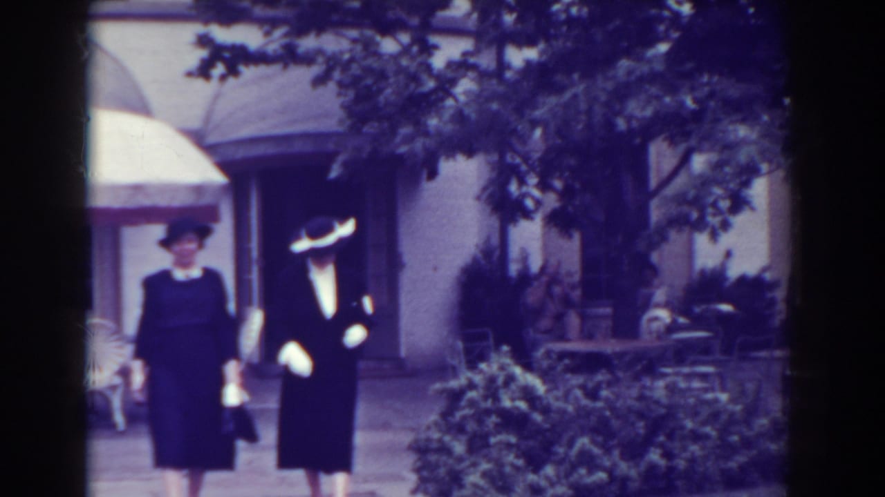 Two women in dark formal clothing stand or walk near a courtyard with shrubs and trees.