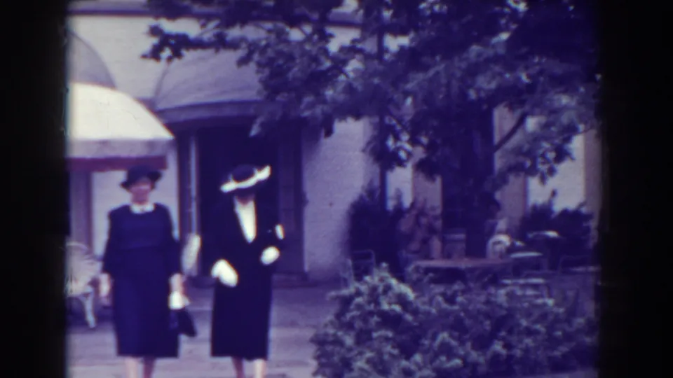 Two women in dark dresses and hats appear closer to camera in the Atlantic City garden setting, with one hat showing a light brim.