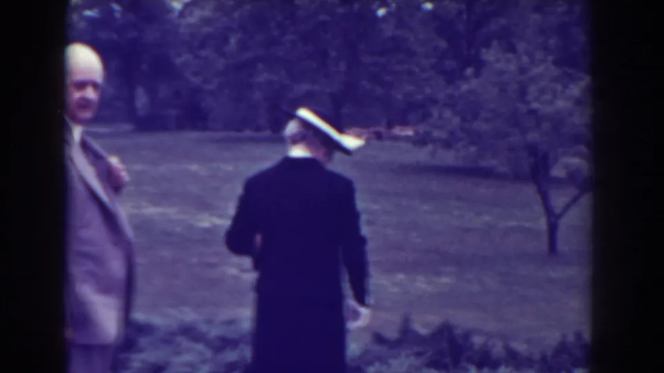 The scene shifts to a grassy area where a man in a light suit faces camera and a woman in dark formal clothing stands with her back turned.