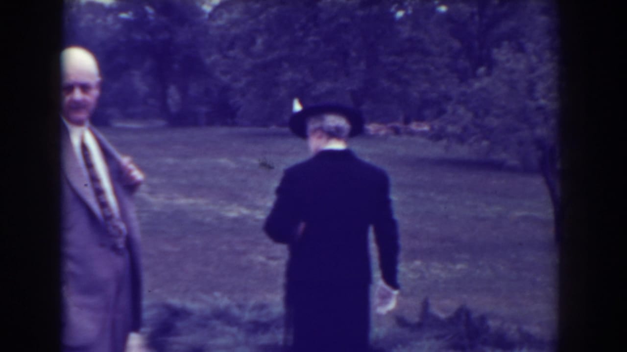 Man in a light suit beside an elderly woman walking away through a garden.