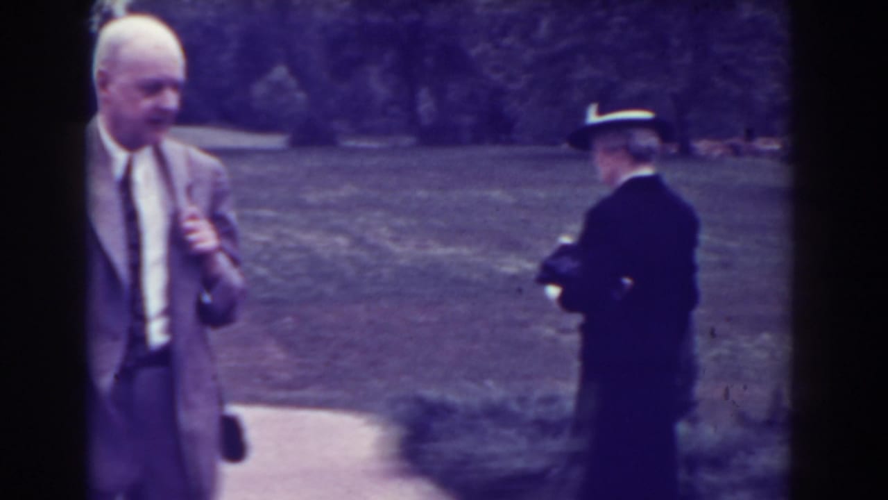 1938 Atlantic City frame with an elderly woman in a dark coat and brimmed hat on a garden path, with a suited man nearby.