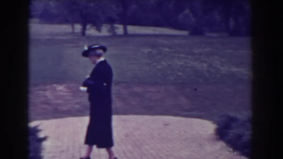 1938 Atlantic City home-movie frame showing an elderly woman in formal dark clothing walking beside a lawn and trees.