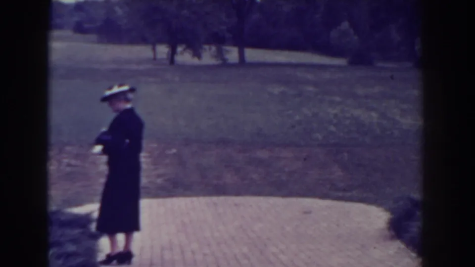 1938 Atlantic City frame showing the elderly woman in a dark coat and hat standing on a paved garden path near lawn and trees.