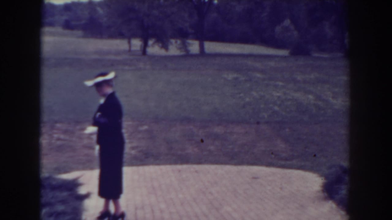 Woman in a dark dress and wide-brimmed hat stands on a paved park walkway.