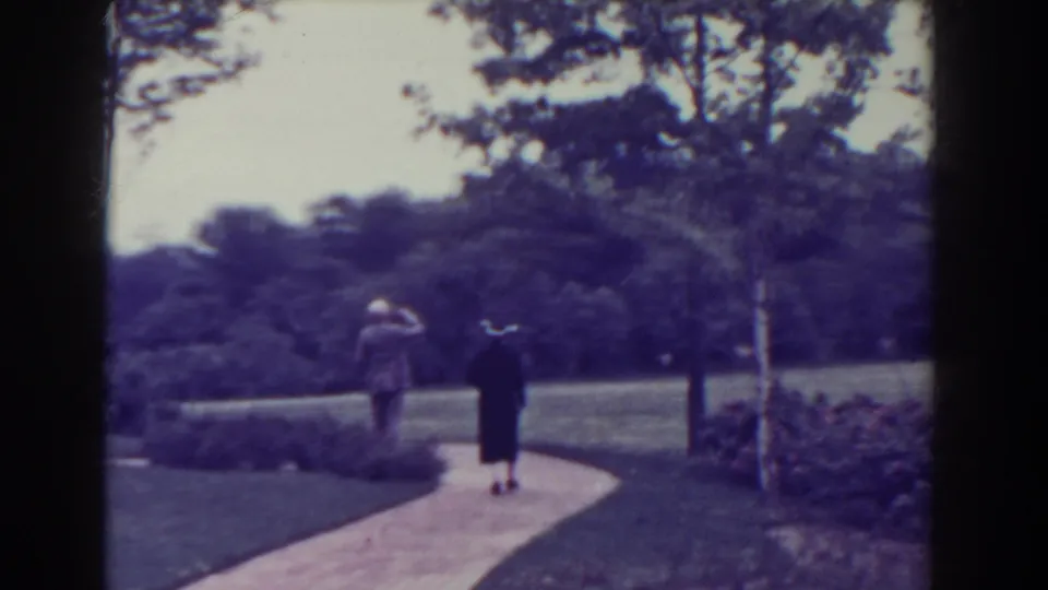 The couple continues down the curved walkway, surrounded by lawn, shrubs, and trees in soft, faded 8mm color.