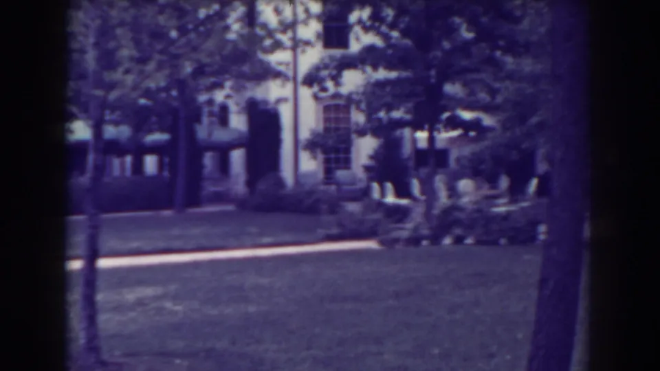 1938 Atlantic City home-movie frame with a house exterior, lawn, path, trees, shrubbery, and outdoor chairs visible.