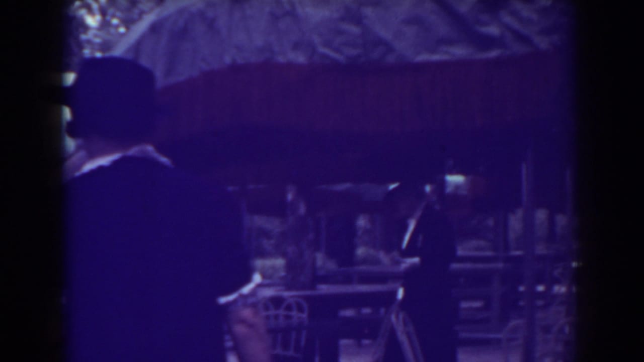 Formally dressed adults in hats stand near outdoor tables and large umbrellas.
