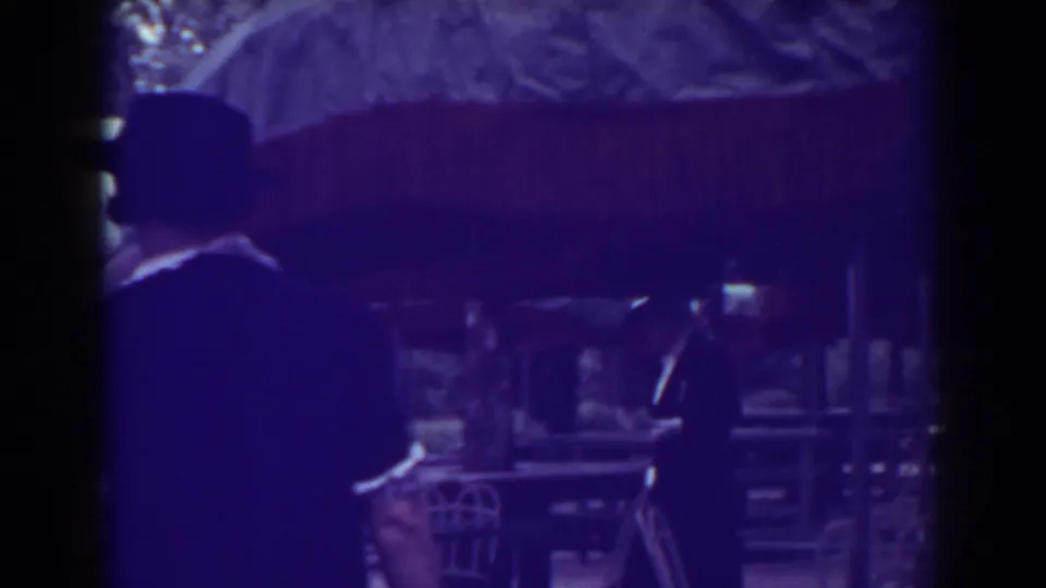 1938 Atlantic City home-movie frame showing adults in dark formal clothing near terrace tables and fringed umbrellas.