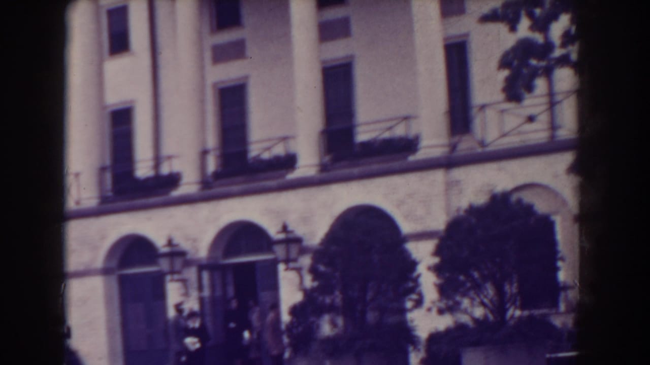 White columned building with arched openings, shrubs, trees, and small figures near the entrance.