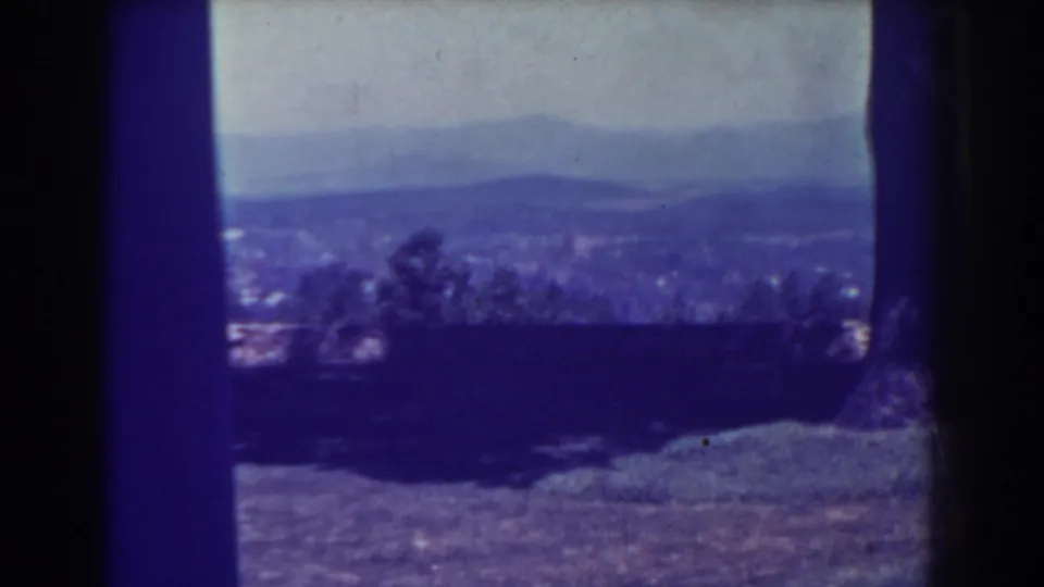 1938 8mm home-movie frame of a distant valley or town and low hills, partially blocked by dark film edges and blue-purple color shift.