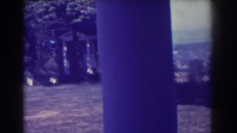 1938 8mm frame with a small house or structure among trees, open ground, distant hills, and heavy blue-purple film cast.