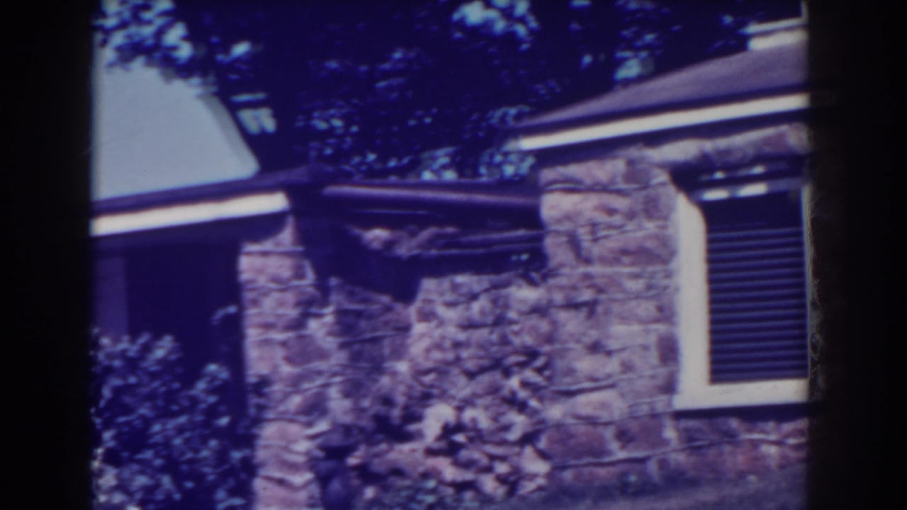 Stone house exterior with shuttered window, roofline, and trees above.