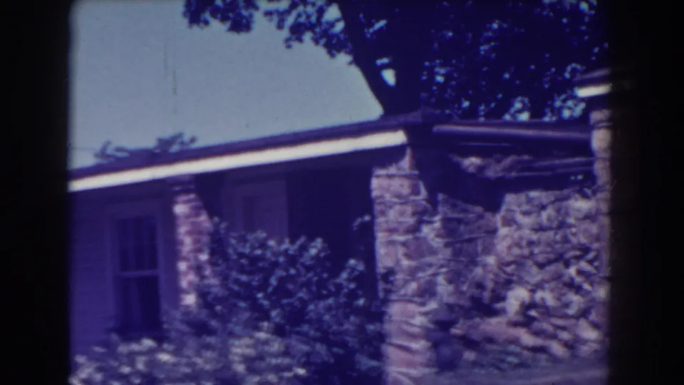 1938 Atlantic City home-movie frame with a tilted view of a house exterior, stone chimney or wall, clapboard siding, windows, and front shrubs.
