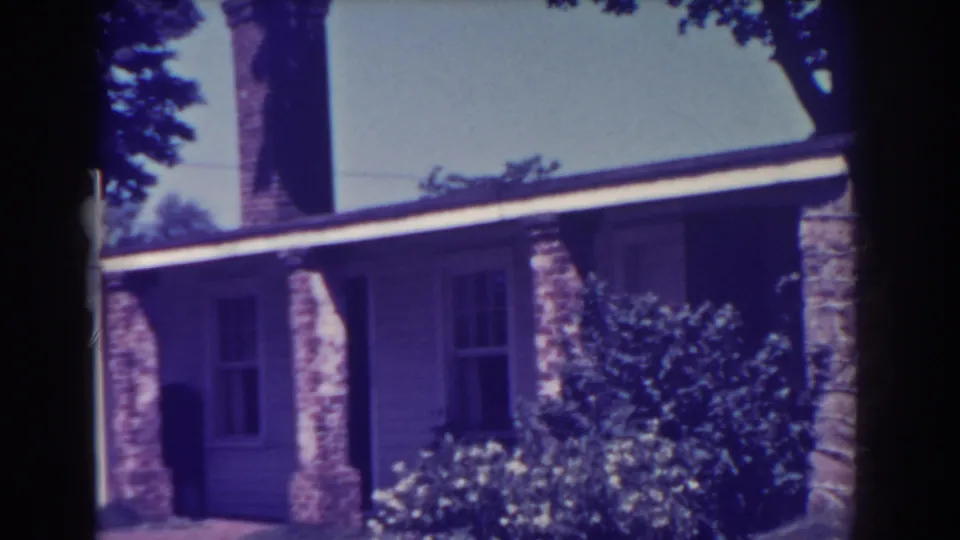 1938 Atlantic City home-movie frame showing a house exterior with a tall chimney, masonry porch supports, clapboard siding, and garden shrubs.