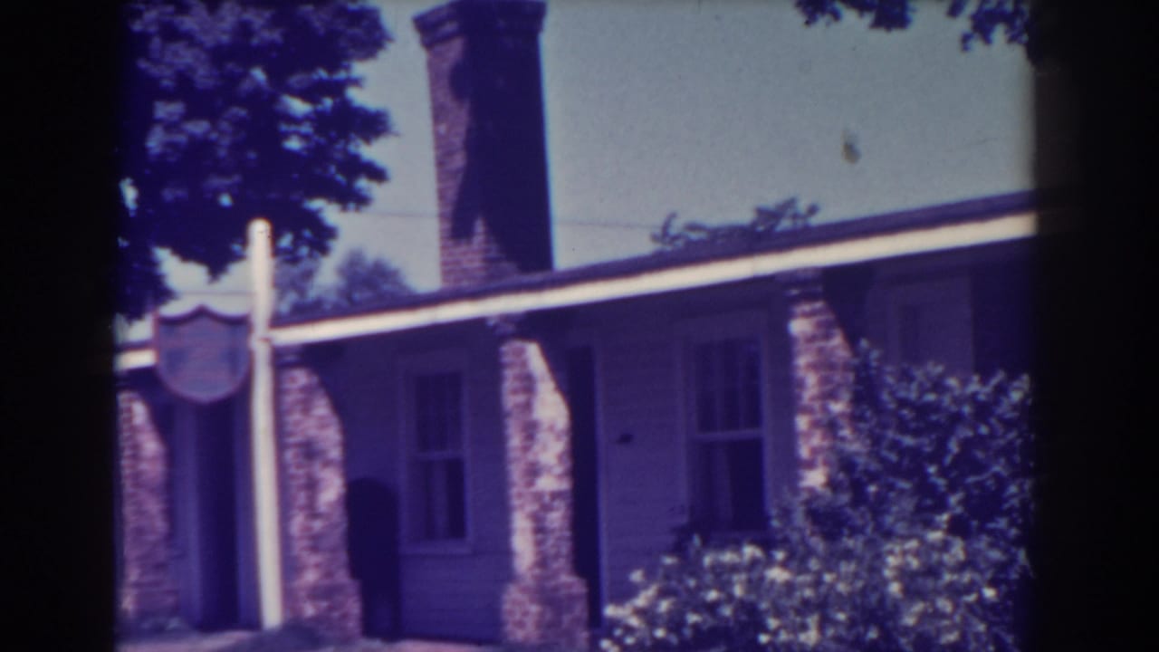 1938 Atlantic City exterior view with a signpost near the entrance, tall chimney, brick porch columns, siding, and dense garden shrubs.