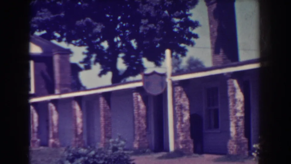 Late frame from the 1938 Atlantic City house exterior, showing the signpost, brick supports, chimney, dark porch opening, and shaded plantings.