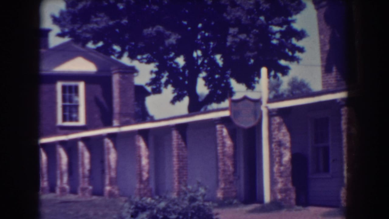 Brick building with a covered pillar-lined walkway, lawn, trees, and a lamp post.