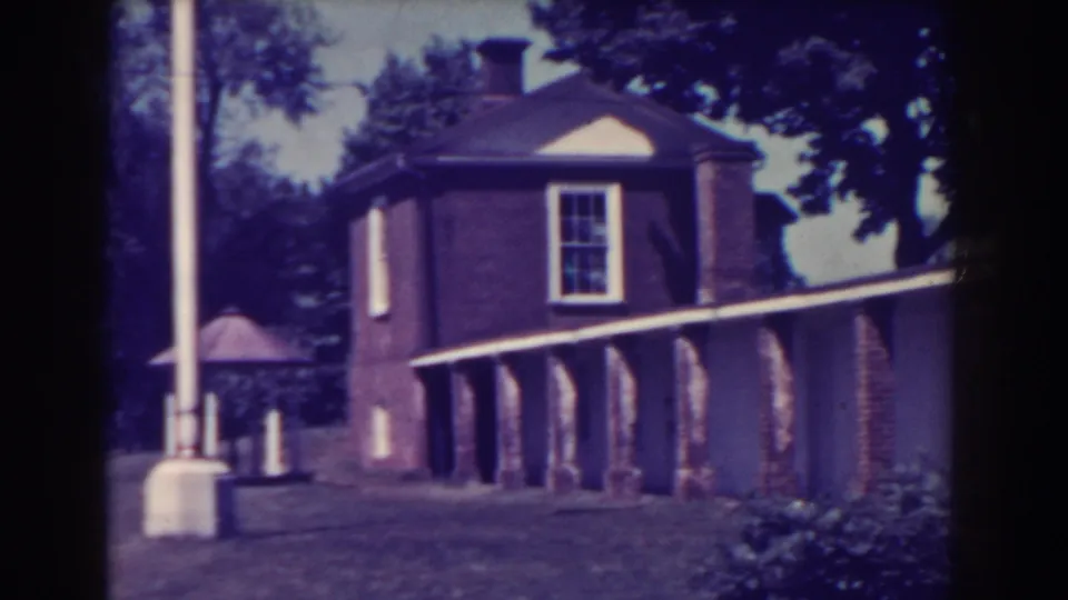 Atlantic City 1938 home-movie frame with a brick building corner, columned walkway, lamp post, grass, shrubs, and trees.