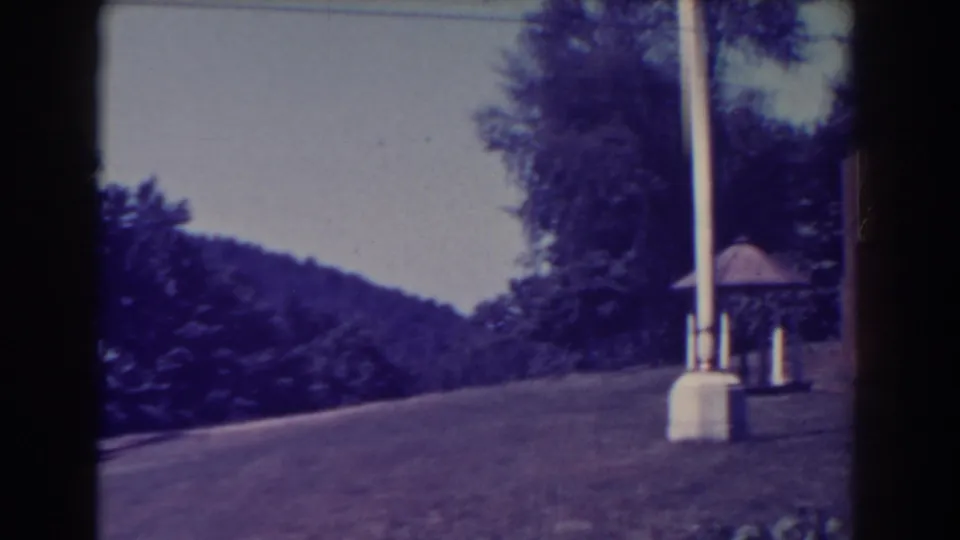 1938 Atlantic City home-movie frame looking across a sloped lawn toward a white pole, small gazebo, trees, and wooded hillside.