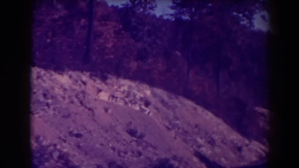 1938 8mm view near Blackwater Falls showing a rocky roadside slope, trees, and a soft sky from a moving vehicle.