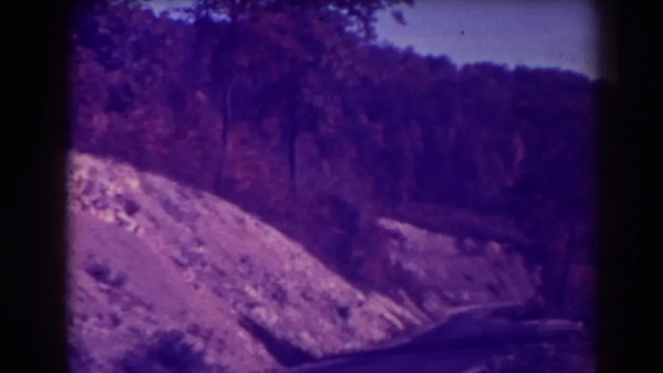 Visitor-shot 1938 8mm frame of a wooded West Virginia roadway with a rocky embankment and dark frame edges.