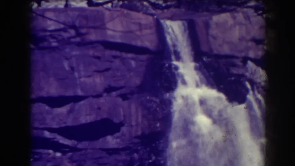 Purple-shifted 1938 8mm frame showing Blackwater Falls pouring over dark rock ledges with bright white water and soft film grain.