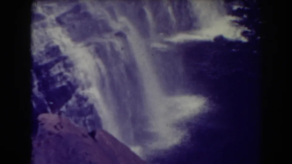 The camera view reaches the lower falls, showing white water, dark water below, and reddish rock in the foreground.