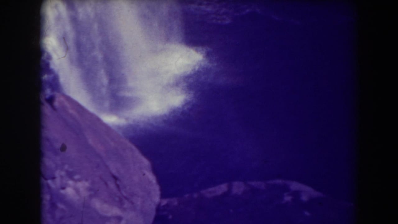 Purple-tinted waterfall and cliff seen from above with dark film borders.