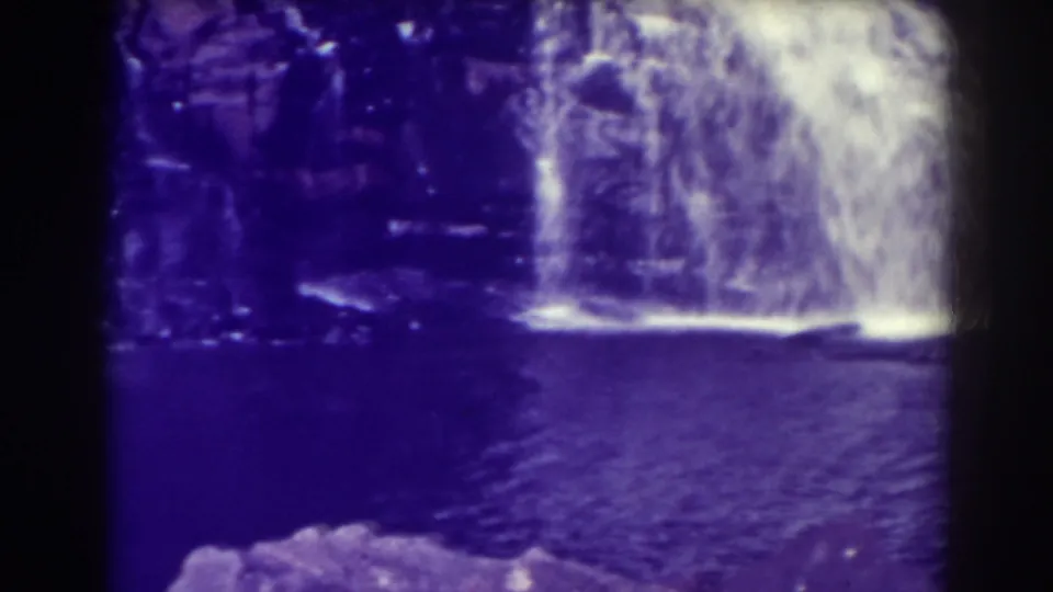 1938 Blackwater Falls home-movie frame showing water spilling over a rocky ledge into a dark pool, with purple color cast and 8mm frame edges.