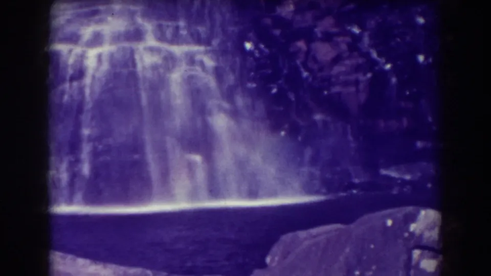 1938 8mm frame of Blackwater Falls with a luminous sheet of water, misty base, dark pool, and surrounding rock ledges.