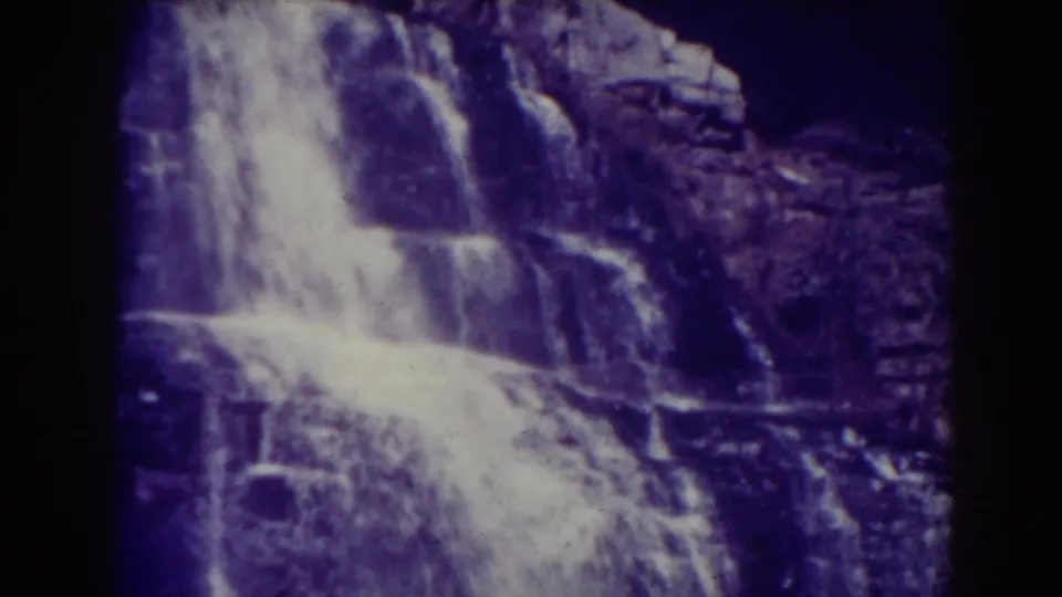 Vintage 8mm frame of Blackwater Falls, with rushing white water spreading across dark rock ledges and soft archival blur.