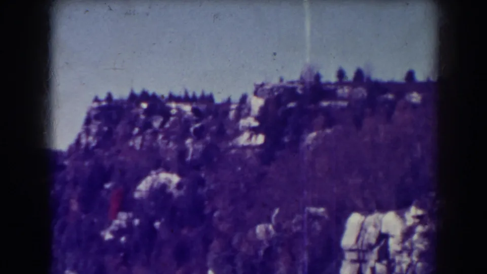 Archival 8mm view of a rocky, tree-covered hillside near Blackwater Falls, with a pale sky and scattered light stone or snow patches.
