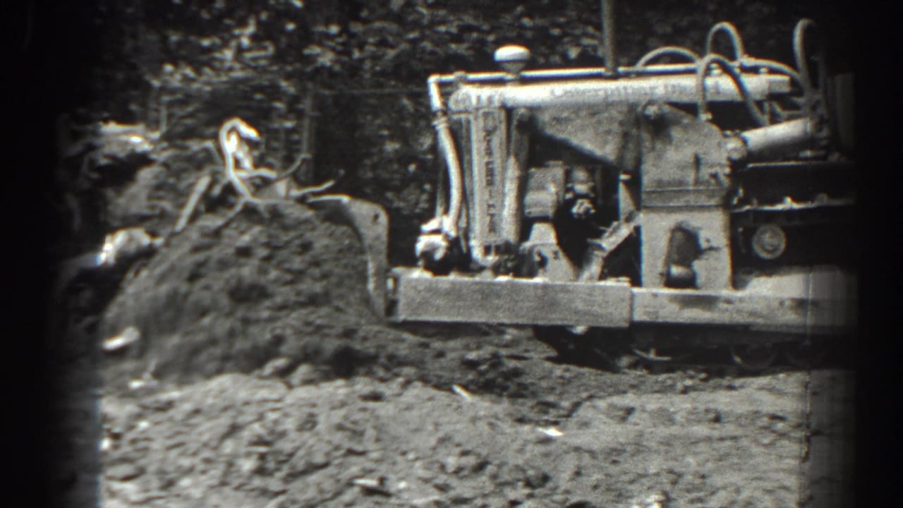 The tractor appears behind churned soil and roots, with its front implement crossing the frame in black-and-white 8mm footage.