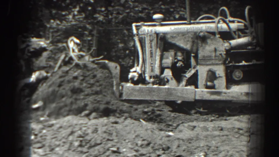 The tractor appears behind churned soil and roots, with its front implement crossing the frame in black-and-white 8mm footage.