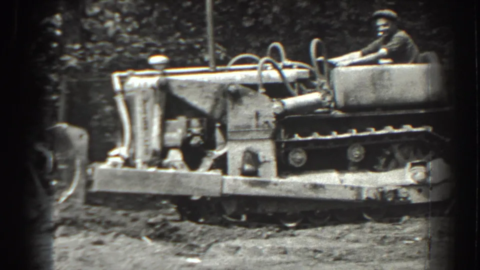 A man operates the tracked tractor in a wooded Cumberland work area, with the machine's tracks, front implement, and churned earth visible.