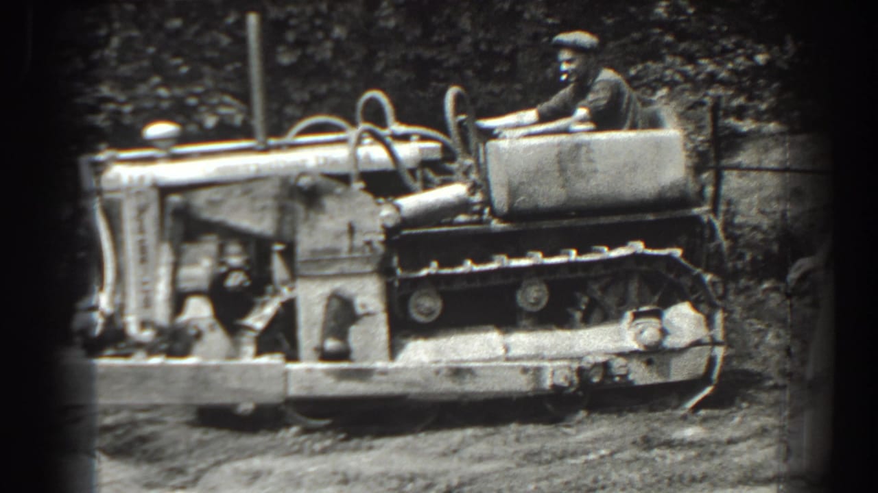 Man in a cap drives a tracked tractor past trees and rough ground.