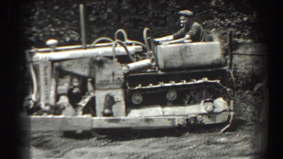 In 1938 Cumberland, Maryland footage, a man in a cap operates a tracked tractor in a wooded outdoor area.