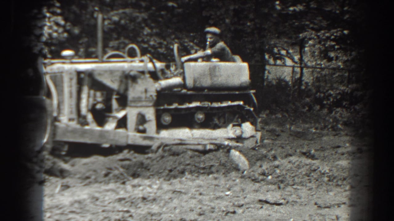 Man seated on a tracked tractor moving over rough dirt near trees and a fence.