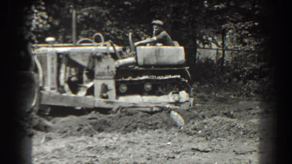 1938 Cumberland, Maryland home-movie frame showing a man operating a tracked tractor across uneven dirt ground near trees and fencing.