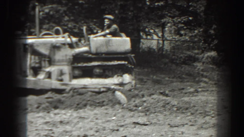 Black-and-white 8mm frame of the tracked tractor in motion, with the operator seated above the machine on rough ground in Cumberland, Maryland.