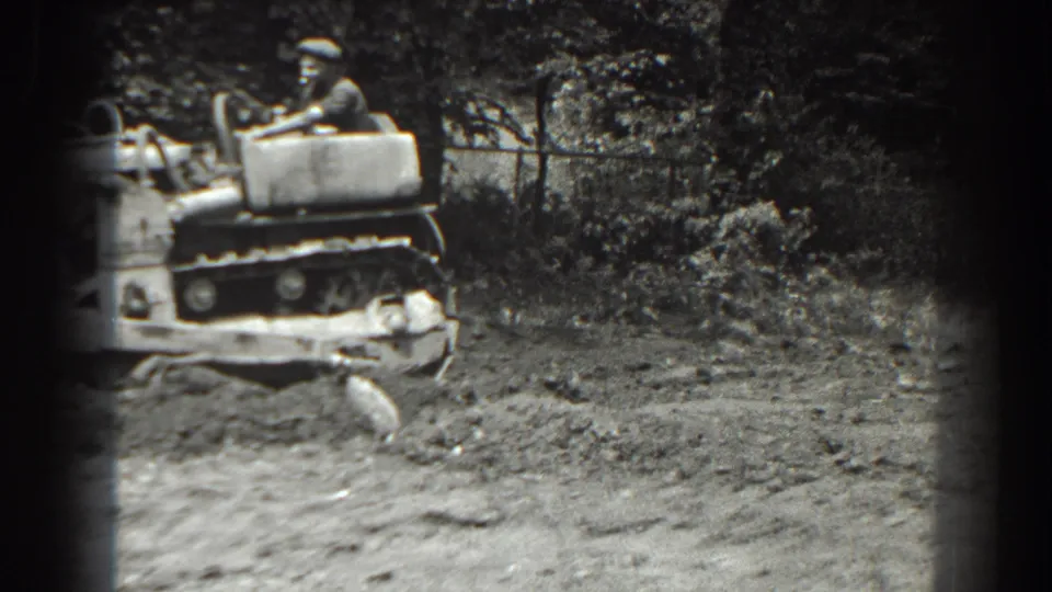 The tractor is partly cropped in the 8mm frame as the operator moves along a rough dirt area bordered by brush and fencing.