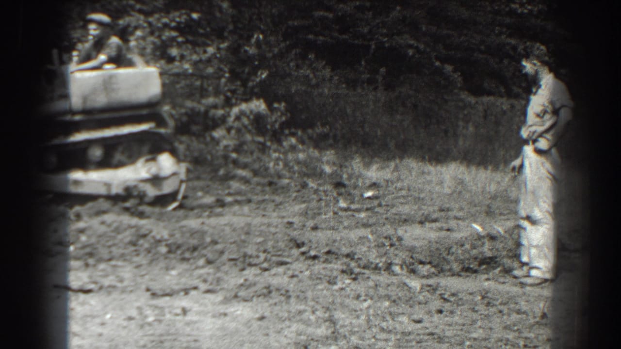 Man in work clothes stands on a dirt work area near a small machine at the left edge.