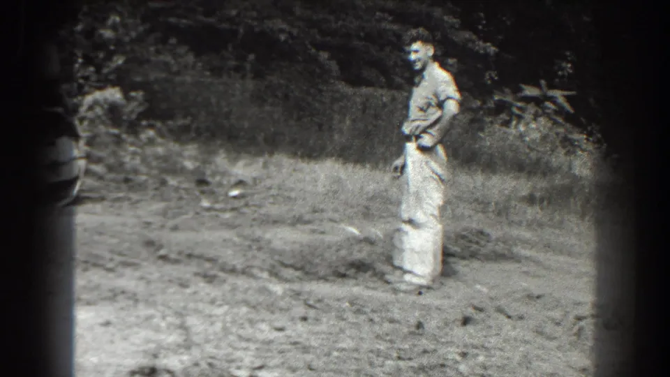 A man in light work clothes stands near a dirt road or work site in the 1938 Cumberland home-movie footage.