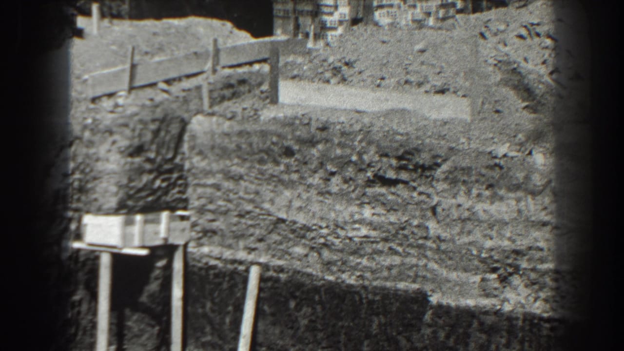 Excavated dirt bank with wood planks, posts, gravel, and distant buildings.