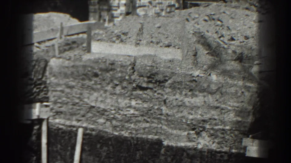 Black-and-white 8mm view of a Cumberland excavation with a timber rail, uneven earth, loose stones, and background buildings.