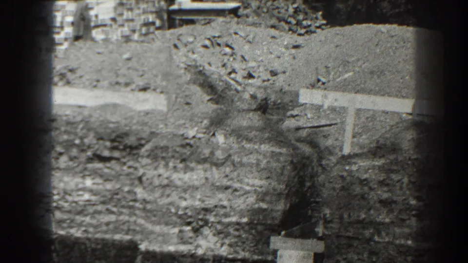 Final 8mm frame of the Cumberland site showing a dirt mound, rough stones, timber rail, and squared foundation edge in bright daylight.