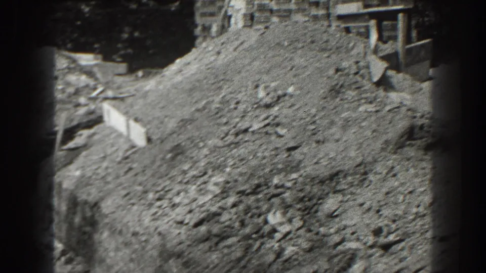 1938 Cumberland home-movie frame showing a dirt pile, rough construction ground, stacked blocks, and part of a nearby building.