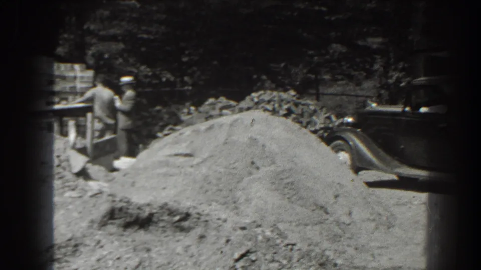 Two men work beside dirt piles and a wooden structure, with a dark old car partly visible at right in the 1938 Cumberland footage.