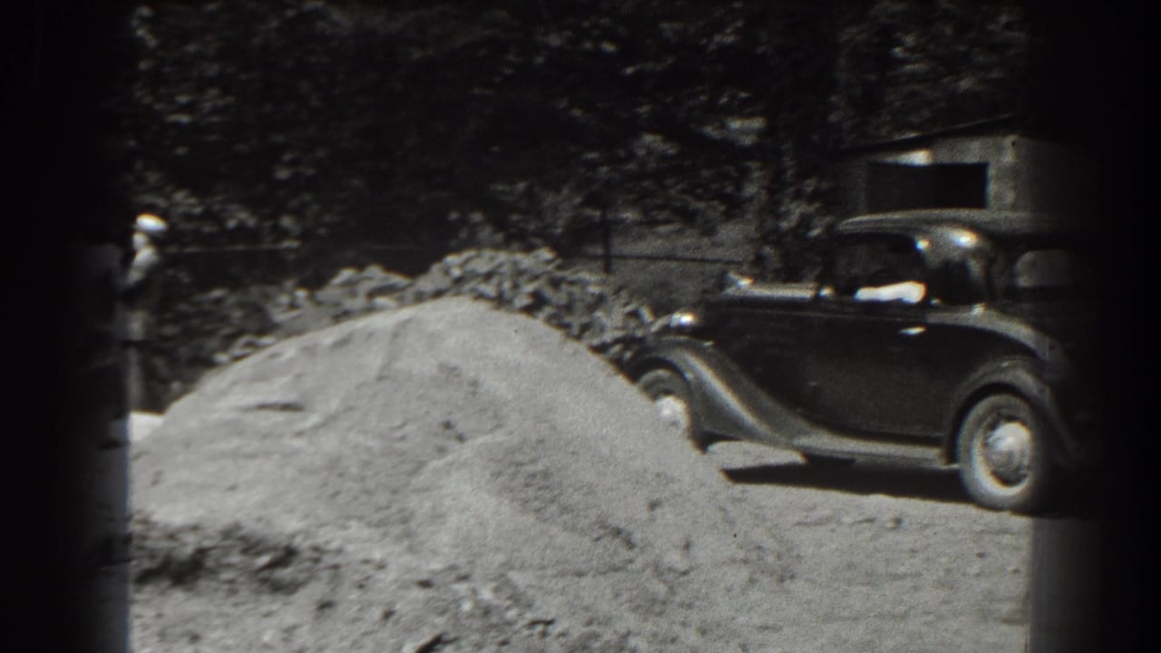 Dark 1930s sedan beside a large sand pile in a wooded outdoor area.