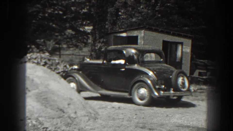 1938 Cumberland, Maryland frame of a rounded 1930s sedan near sand, rocks, trees, and a small shed.